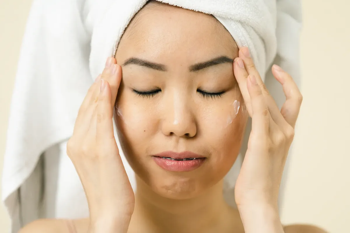 Asian woman with a towel on head applying moisturizer for skincare routine.