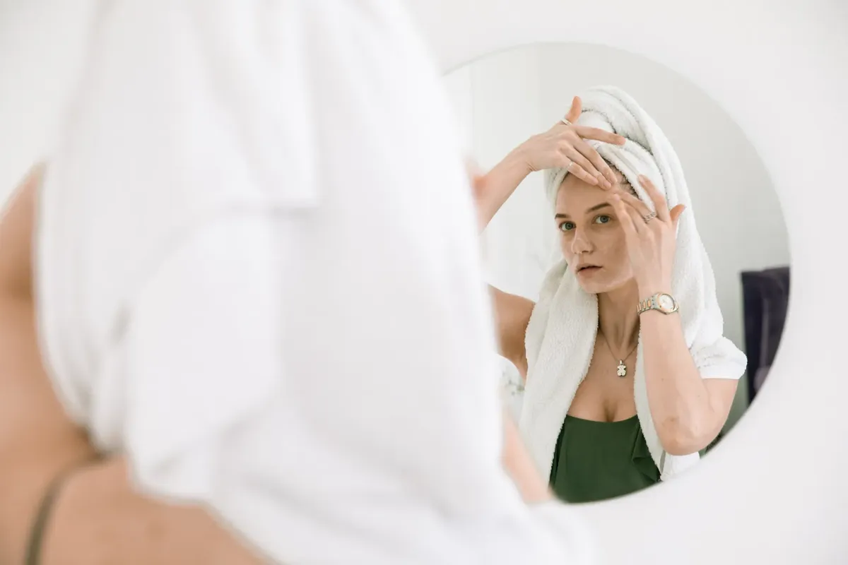 Woman with towel on head applying skincare, enjoying beauty routine in front of mirror.
