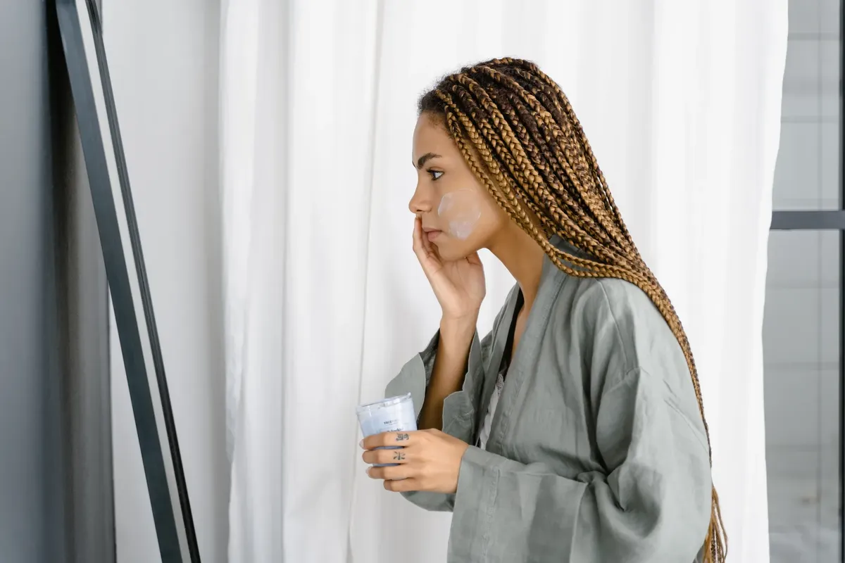 Young woman in robe applying moisturizer in front of mirror as part of her daily skincare routine.