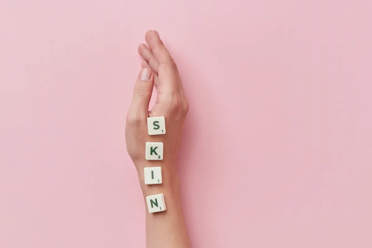 A creative arrangement of Scrabble tiles spelling 'skin' on a hand against a pink backdrop.